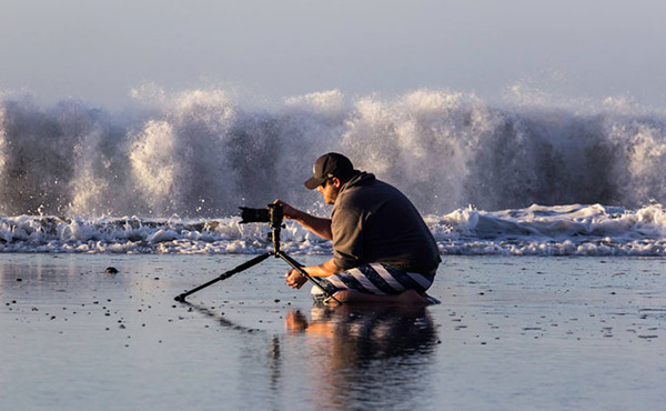 Jay Olson knelling in the surf taking a photo of the sunrise while a large wave crashes in the background