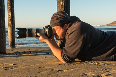 Jay Olson laying on his stomach in the sand holding his camera to his eye while taking a photo of the beach