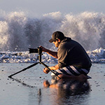 Jay Olson kneeling in the surf taking a photo of the sunrise while a large wave crashes in the background